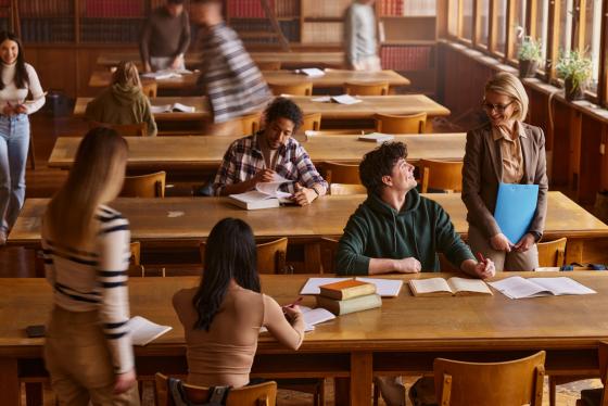 Blid, kvinnelig professor snakker med mannlig student i biblioteket med flere studenter rundt