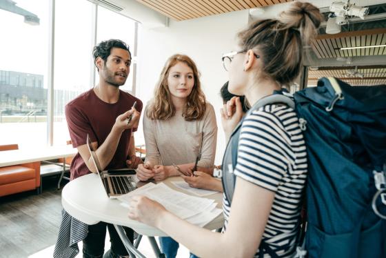 Tre studenter st&aring;r ved et bord og diskuterer