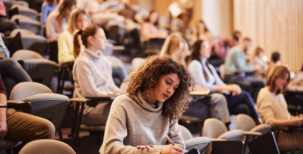 En kvinnelig student sitter i et auditorium og skriver med mange medstudenter bak seg