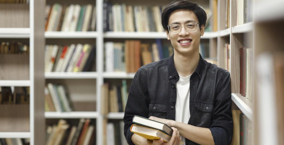 Smilende mannlig student holder en bokstabel i biblioteket
