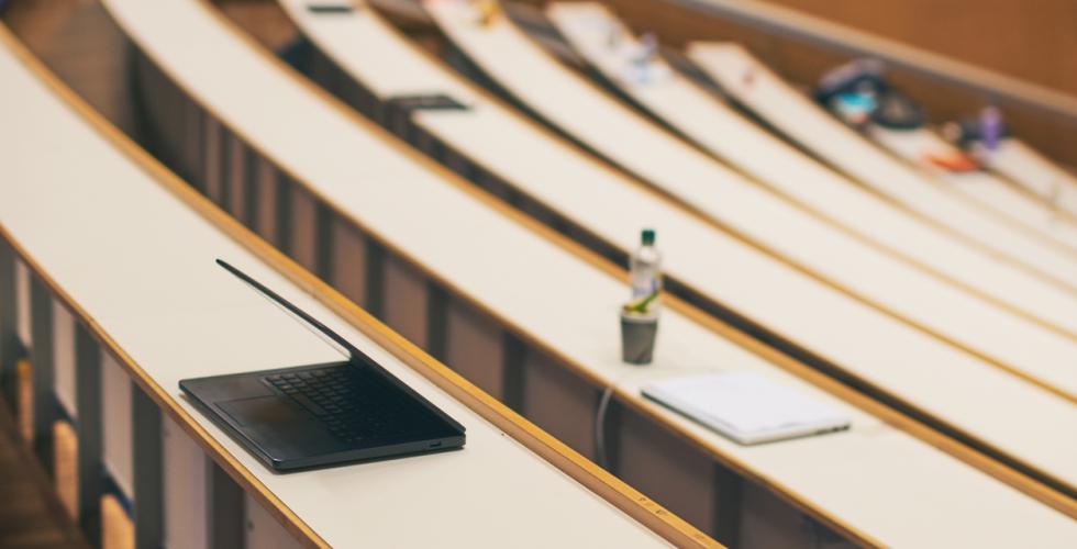 Én laptop på pulten blant mange tomme pulter i et tomt auditorium på universitetet
