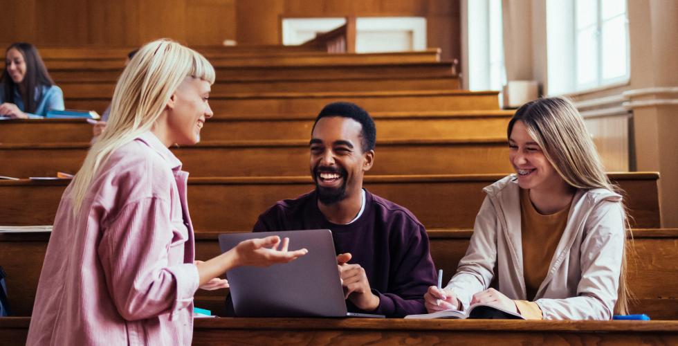 Tre studenter sitter i et auditorium og diskuterer smilende