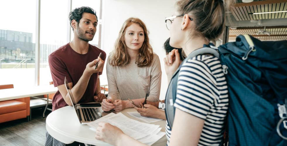 Tre studenter st&aring;r ved et bord og diskuterer